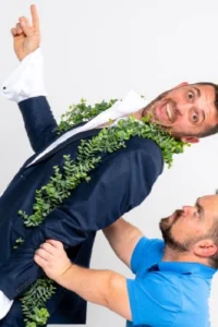 Dvojica muškaraca u promotivnoj fotografiji za predstavu Ljudina: jedan u tamnoplavom odijelu ovijen zelenim vijencem podignut je u zrak s izrazom iznenađenja na licu, drugi u plavoj polo majici ga pridržava i gleda prema gore, bijela pozadina