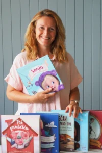 Ilustratorica Nikolina Novosel Nežmah u ružičastoj bluzi drži slikovnicu Sanja uvijek ima vremena, ispred nje poredane dječje knjige koje je ilustrirala