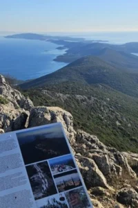 Informativna ploča na kamenitom vrhu s pogledom na pošumljeni greben i razvedenu obalu otoka pod plavim morem.