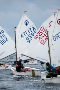 Mladi jedriličari u jedrilicama klase Optimist plove oko velike narančaste plutače na valovitom moru, s bijelim jedrima i oznakama CRO i ITA.