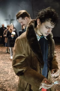 A scene from Drvene ptice with a man in a brown coat in the foreground and a line of actors behind him, with a bare tree and a cross in the background.