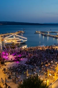 Večernji pogled iz zraka na crikveničku rivu: trg ispunjen publikom ispred pozornice osvijetljene ljubičastim svjetlom, lijevo osvijetljeni panoramski kotač, uz obalu osvijetljeni brodovi i molovi, a u pozadini more i obala u sumrak
