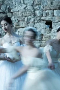 Ballerinas in long white ballet dresses in front of a stone wall, with some of the dancers blurred in motion.