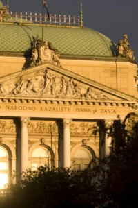 Pročelje HNK-a Ivana pl. Zajca u Rijeci obasjano toplim svjetlom, sa stupovima, reljefima i zelenim krovom.