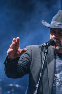 Riccardo Staraj u tamnom šeširu i sakou nastupa pred mikrofonom, gestikulirajući rukama pod plavim scenskim svjetlom.