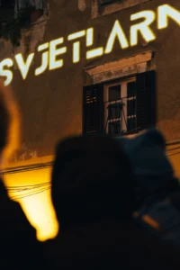 Osvijetljeni natpis Svjetlarna projiciran na staru kamenu fasadu s prozorom i škurama, dok posjetitelji u prvom planu snimaju prizor.
