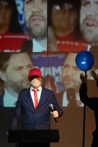 Scenski prizor s izvođačem prekrivena lica u odijelu, crvenoj kravati i kapi te figurom koja podiže plavi balon ispred višestrukih videoprojekcija.