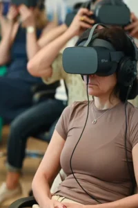 Žena sjedi s VR naočalama i slušalicama, dok drugi gledatelji iza nje namještaju svoje VR naočale.