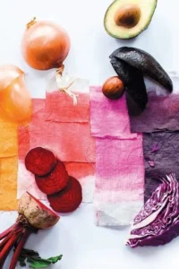 Colorful fabric swatches beside onions, avocado, beetroot, purple cabbage, and spinach on a white surface.