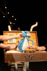 Two performers dressed in black operate a blue-and-white puppet in an open wooden suitcase onstage.
